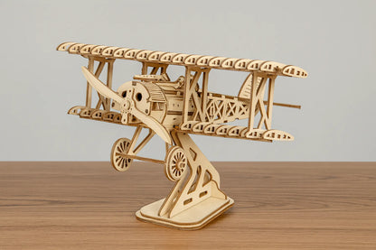 Maquette en bois d’un avion biplan vintage avec hélice et structure ajourée, posée sur une table en bois devant un mur gris clair.
