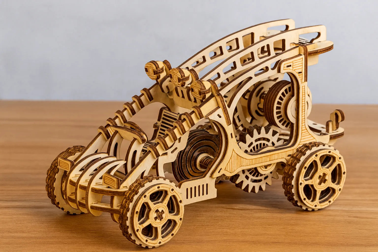 Maquette en bois d’un buggy mécanique, aux grandes roues crantées et aux engrenages apparents, exposée sur une table en bois devant un mur gris clair.