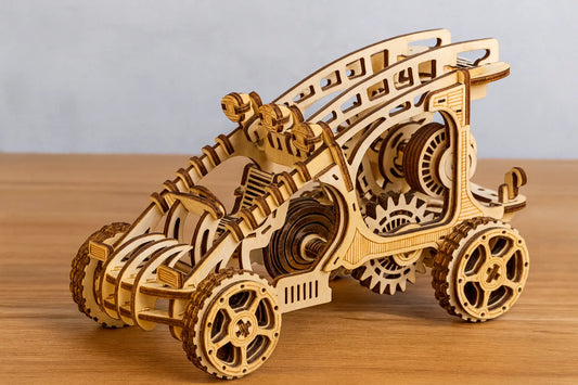 Maquette en bois d’un buggy mécanique, aux grandes roues crantées et aux engrenages apparents, exposée sur une table en bois devant un mur gris clair.