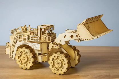 Maquette en bois d’un bulldozer détaillé avec grande pelle avant articulée et pneus crantés, posée sur une table en bois devant un mur gris clair.