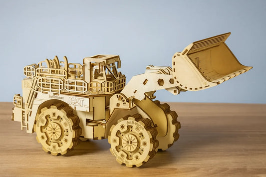 Maquette en bois d’un bulldozer détaillé avec grande pelle avant articulée et pneus crantés, posée sur une table en bois devant un mur gris clair.