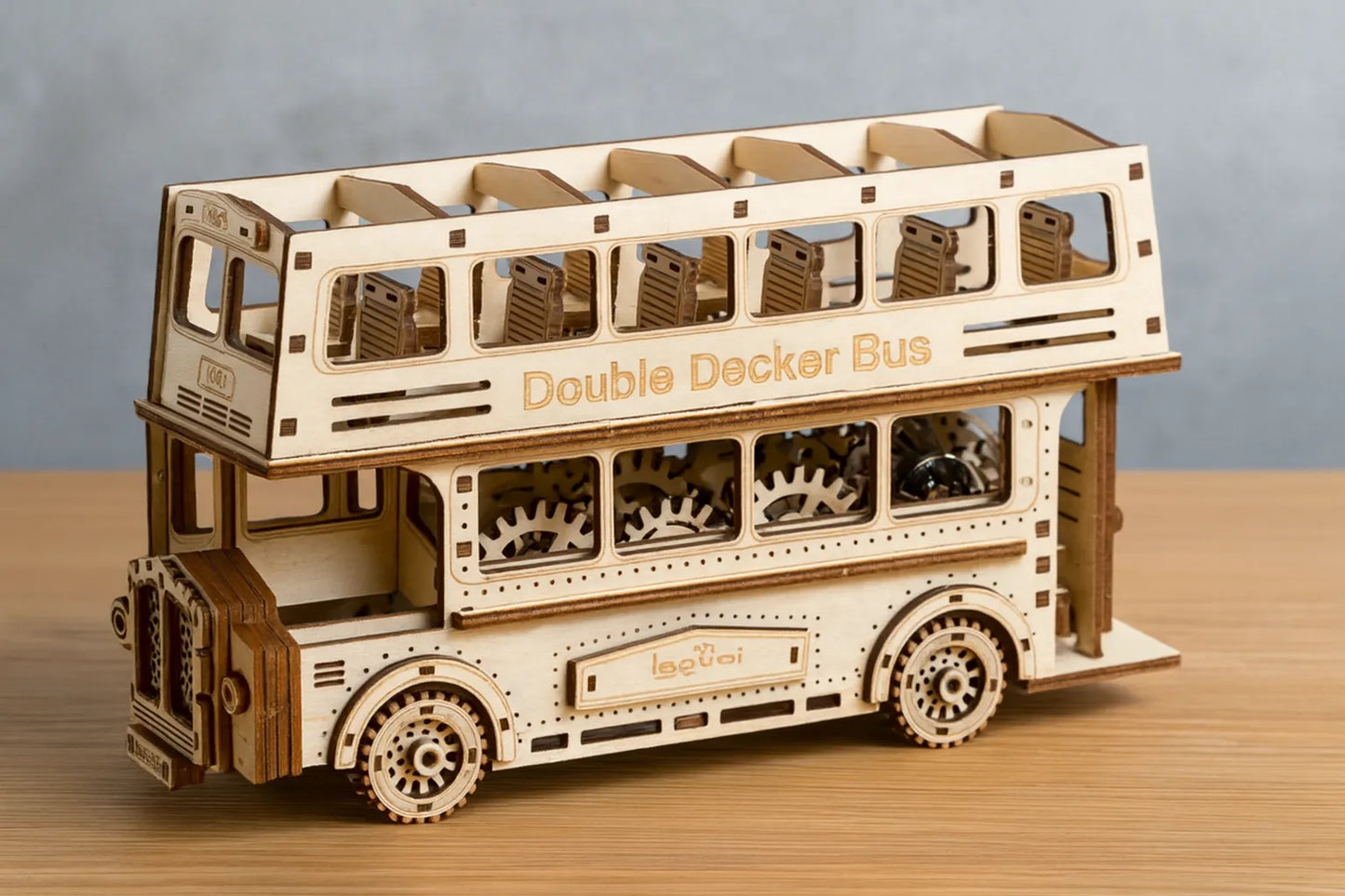 Maquette en bois d’un bus à impériale, à deux étages avec sièges détaillés et engrenages visibles, placée sur une table en bois devant un mur gris clair.