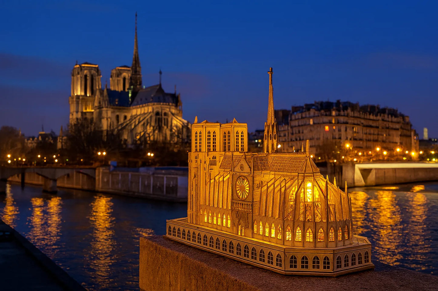 Maquette en bois illuminée de la cathédrale Notre-Dame de Paris posée sur un muret au bord de la Seine, avec la véritable cathédrale en arrière-plan au crépuscule.