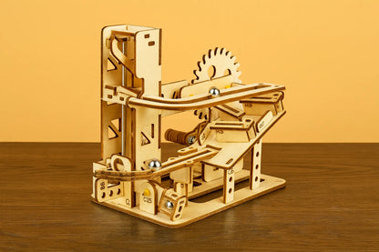 Maquette en bois d’un circuit de billes à rampe, équipée de roues dentées et de rails en spirale, posée sur une table en bois devant un mur orangé.