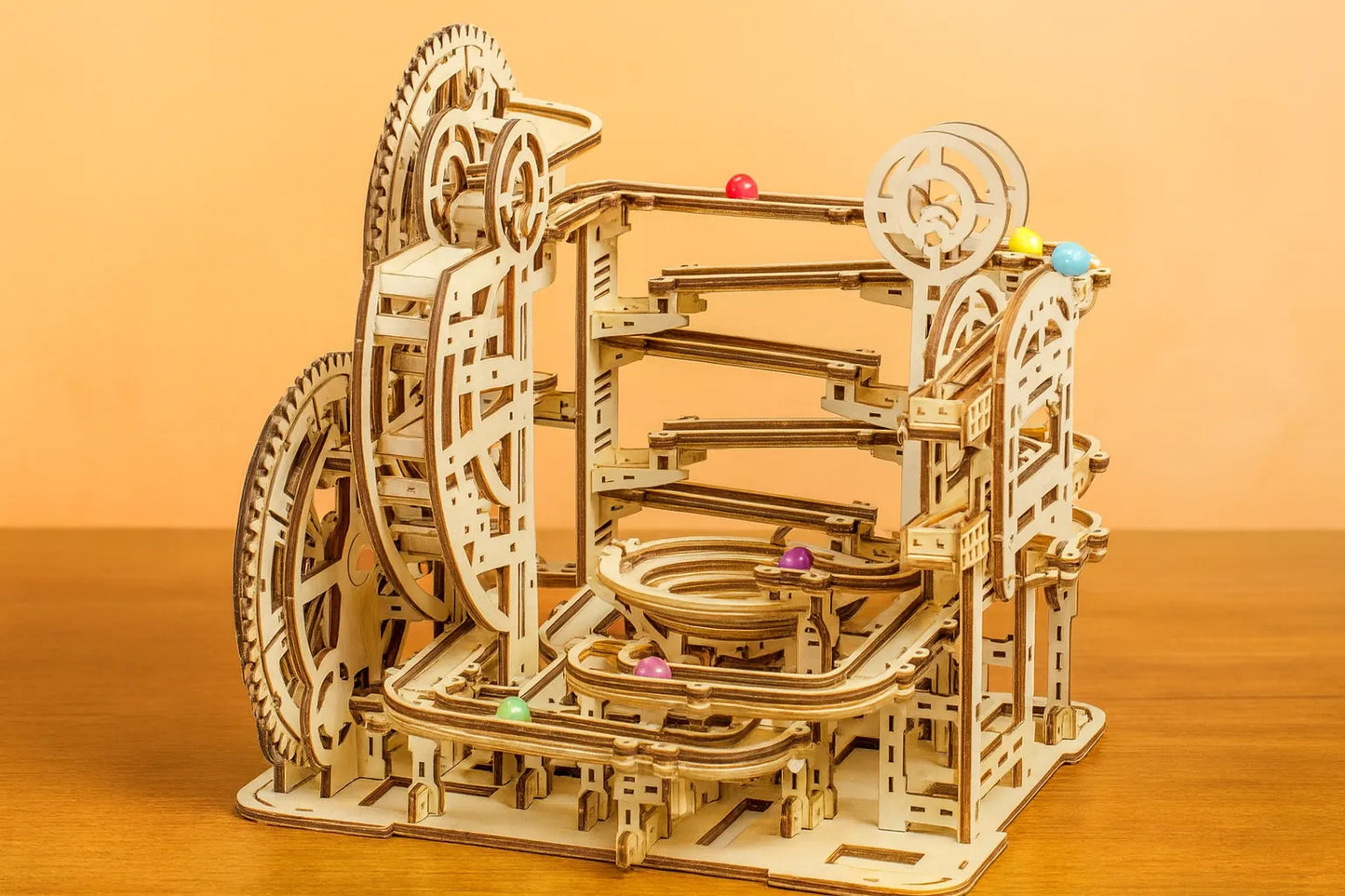 Maquette en bois d’un circuit de billes, formée de multiples rampes et spirales avec des billes multicolores, posée sur une table en bois devant un mur orangé.