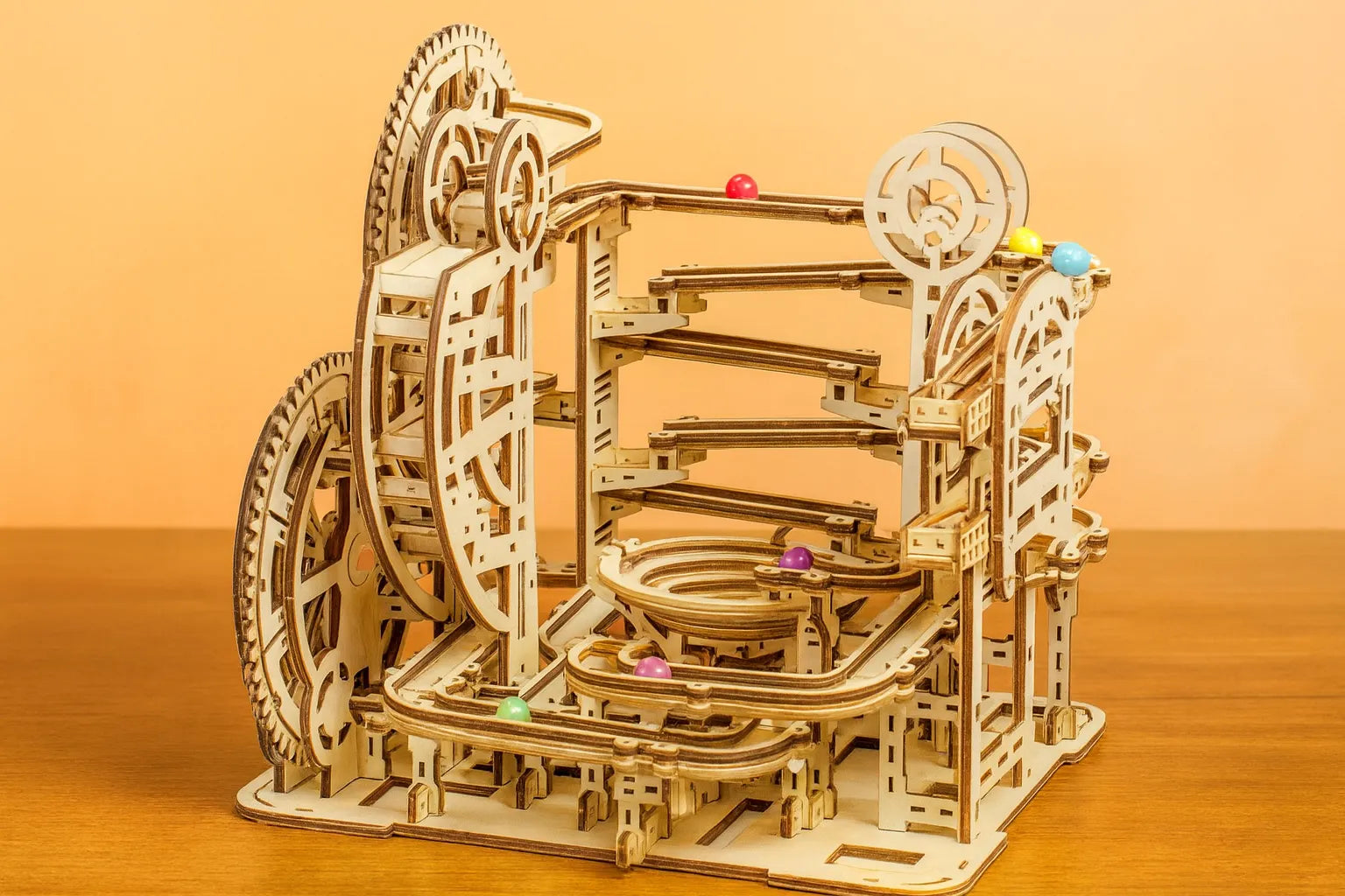 Maquette en bois d’un circuit de billes, formée de multiples rampes et spirales avec des billes multicolores, posée sur une table en bois devant un mur orangé.