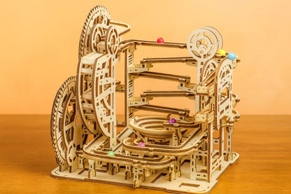 Maquette en bois d’un circuit de billes, formée de multiples rampes et spirales avec des billes multicolores, posée sur une table en bois devant un mur orangé.