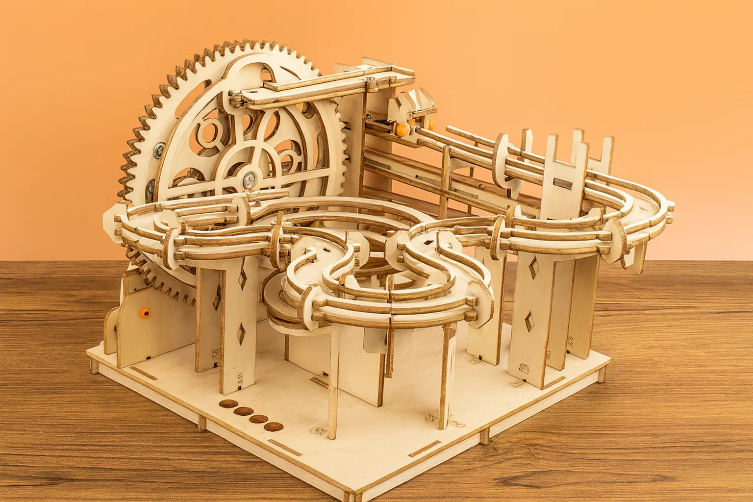 Maquette en bois d’un circuit de billes en zigzag, dotée d’un grand engrenage et de rails courbés, installée sur une table en bois devant un mur orangé.