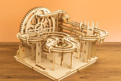 Maquette en bois d’un circuit de billes en zigzag, dotée d’un grand engrenage et de rails courbés, installée sur une table en bois devant un mur orangé.