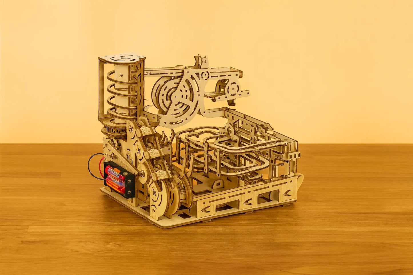 Maquette en bois d’un circuit de billes avec de grands engrenages, disposée sur une table en bois devant un mur orangé.
