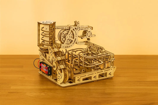 Maquette en bois d’un circuit de billes avec de grands engrenages, disposée sur une table en bois devant un mur orangé.