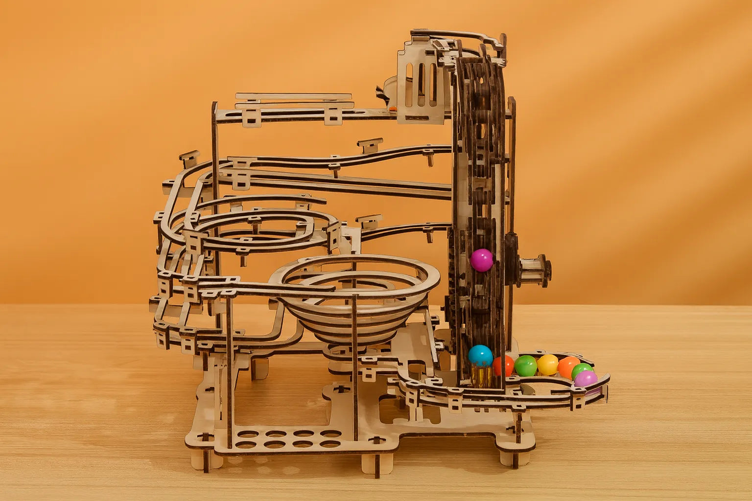 Maquette en bois d’un circuit de billes, composée de spirales, de rails et d’engrenages, exposée sur une table en bois devant un mur orangé.