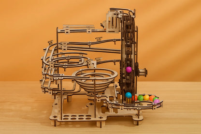 Maquette en bois d’un circuit de billes, composée de spirales, de rails et d’engrenages, exposée sur une table en bois devant un mur orangé.