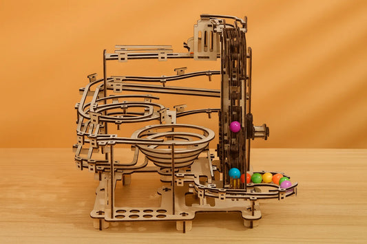 Maquette en bois d’un circuit de billes, composée de spirales, de rails et d’engrenages, exposée sur une table en bois devant un mur orangé.