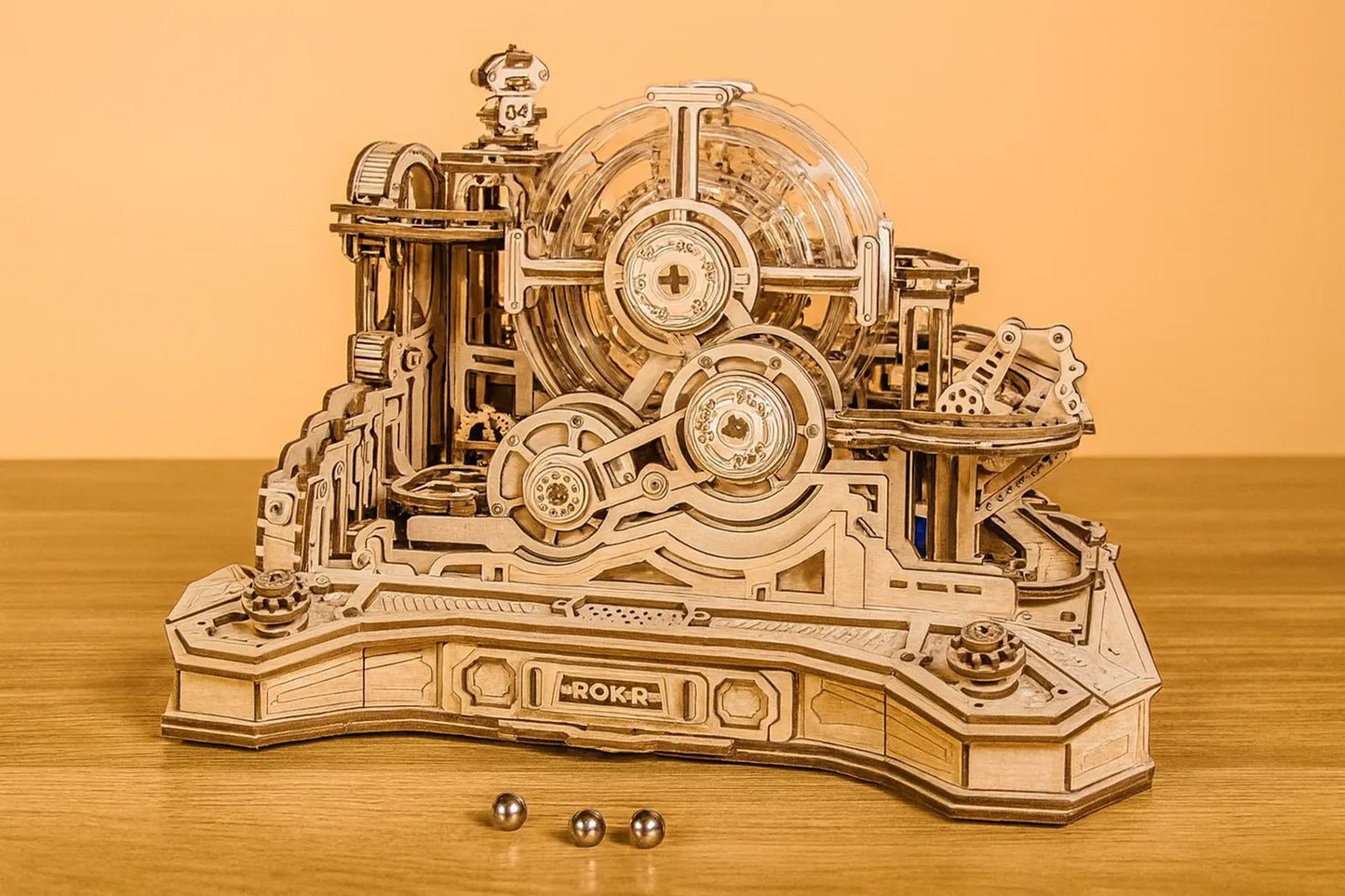 Maquette en bois d’un circuit de billes à orbite mécanique, composée d’engrenages et de rampes, installée sur une table en bois devant un mur orangé.