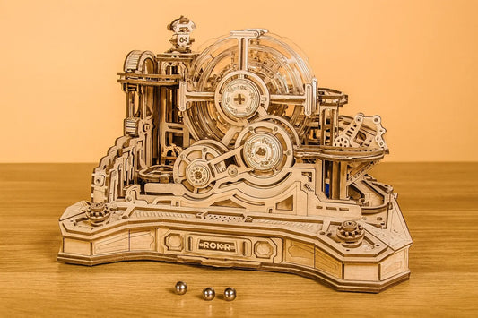 Maquette en bois d’un circuit de billes à orbite mécanique, composée d’engrenages et de rampes, installée sur une table en bois devant un mur orangé.