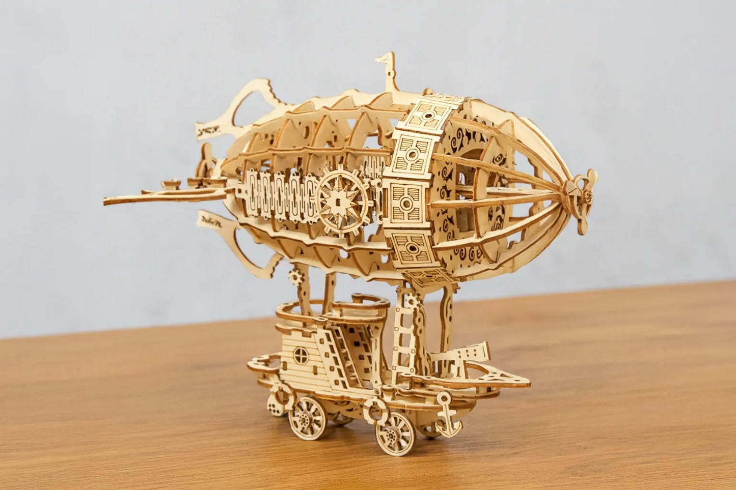 Maquette en bois d’un dirigeable steampunk ajouré, composée d’engrenages et de structures mécaniques, posée sur une table en bois devant un mur gris clair.