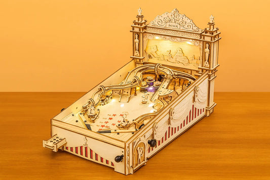 Maquette en bois d’un flipper rétro avec décorations sculptées et lumières intégrées, posée sur une table devant un mur orangé.