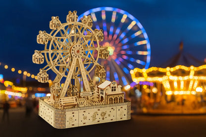 Maquette en bois d’une grande roue lumineuse posée sur une table en bois avec un manège et des guirlandes éclairées à l’arrière.