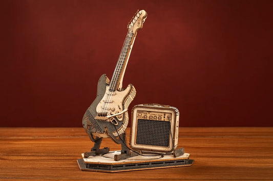 Maquette en bois d’une guitare électrique stylisée accompagnée d’un ampli, posée sur une table en bois devant un fond rouge profond.