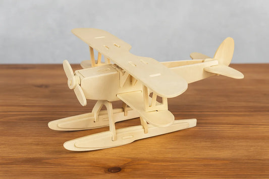 Maquette en bois d’un hydravion biplan clair, équipée de flotteurs, posée sur une table en bois devant un mur gris clair.