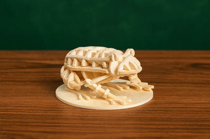 Maquette en bois d’une grenouille, posée sur une table en bois devant un fond vert foncé.