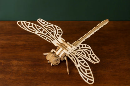 Maquette en bois d’une libellule posée sur une table en bois, grandes ailes ajourées devant un fond vert.