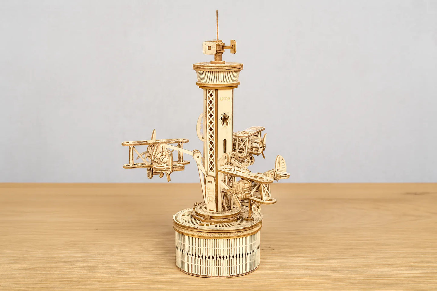 Maquette en bois d’un manège aérien avec de petits avions fixés autour d’une tour posée sur une table en bois devant un mur gris clair.