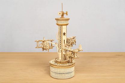 Maquette en bois d’un manège aérien avec de petits avions fixés autour d’une tour posée sur une table en bois devant un mur gris clair.