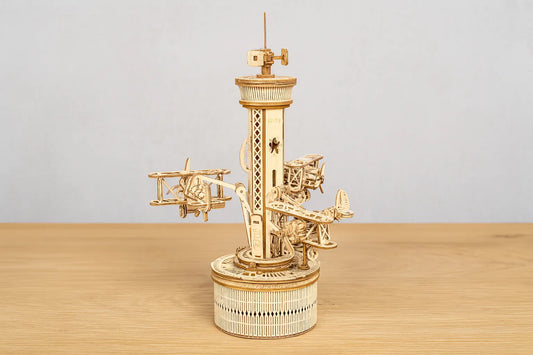 Maquette en bois d’un manège aérien avec de petits avions fixés autour d’une tour posée sur une table en bois devant un mur gris clair.