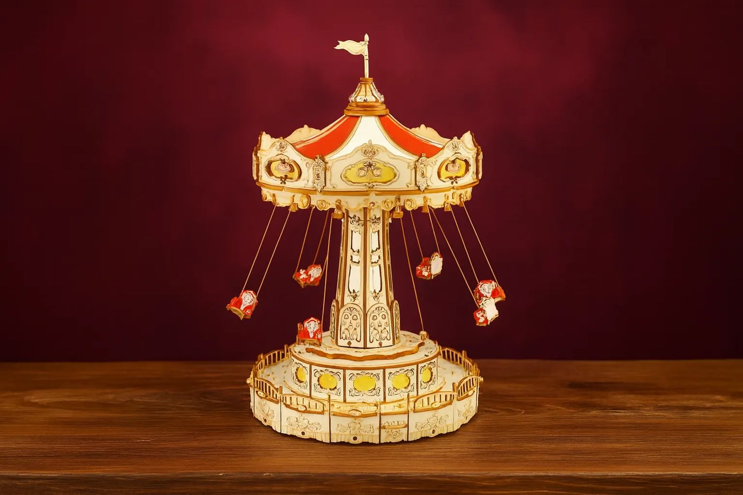 Maquette en bois d’un manège de chaises volantes rouges posée sur une table en bois devant un mur rouge.