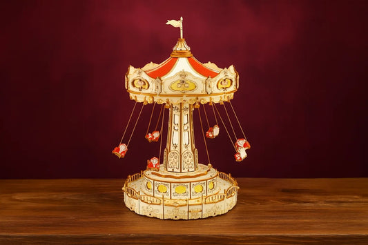 Maquette en bois d’un manège de chaises volantes rouges posée sur une table en bois devant un mur rouge.