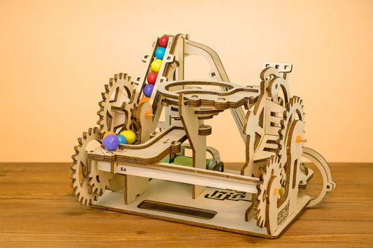 Maquette en bois d’une montée de billes colorées, avec des rampes en boucle, présentée sur une table en bois devant un mur orangé.