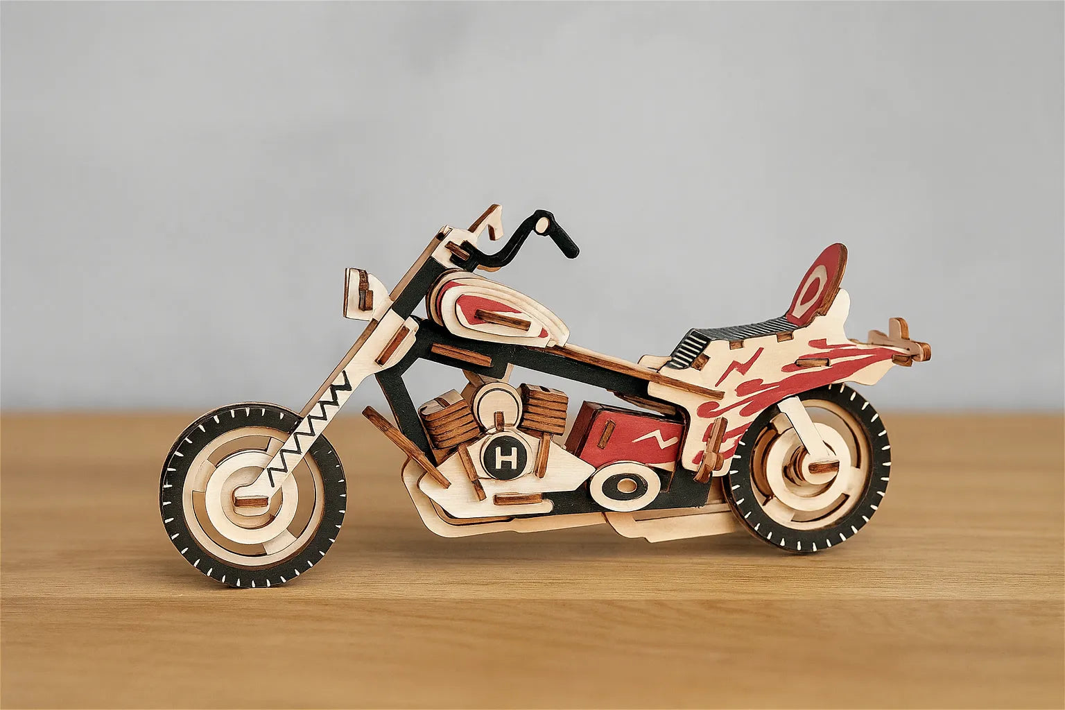 Maquette en bois d’une moto colorée rouge et noire posée sur une table en bois devant un mur gris clair.