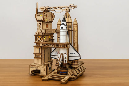 Maquette en bois d’une navette spatiale blanche sur une rampe de lancement en bois, posée sur une table en bois devant un mur gris clair.