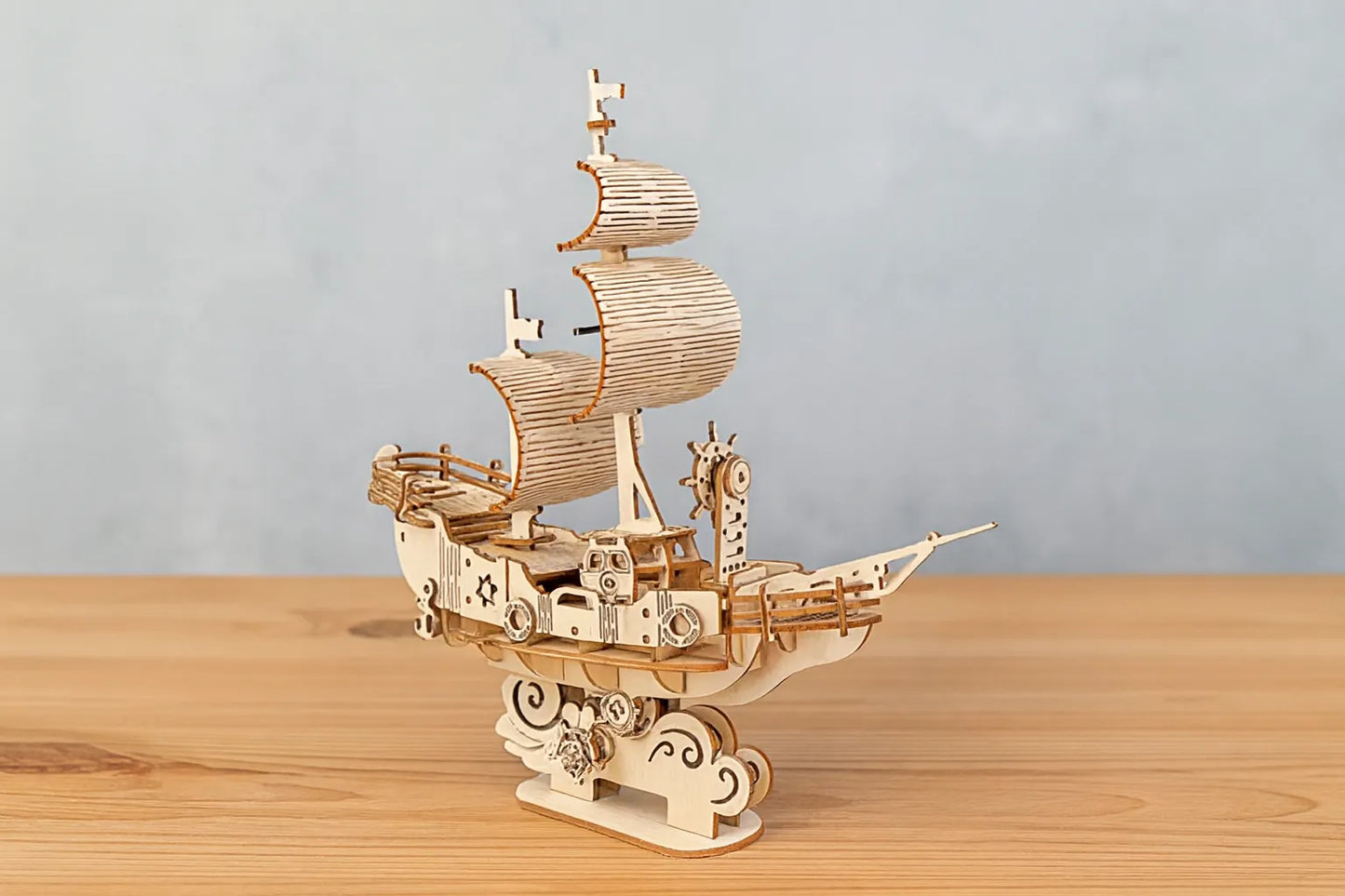 Maquette en bois d’un navire des vents stylisé, ornée de voiles courbées et de roues décoratives, posée sur une table en bois devant un mur gris clair.