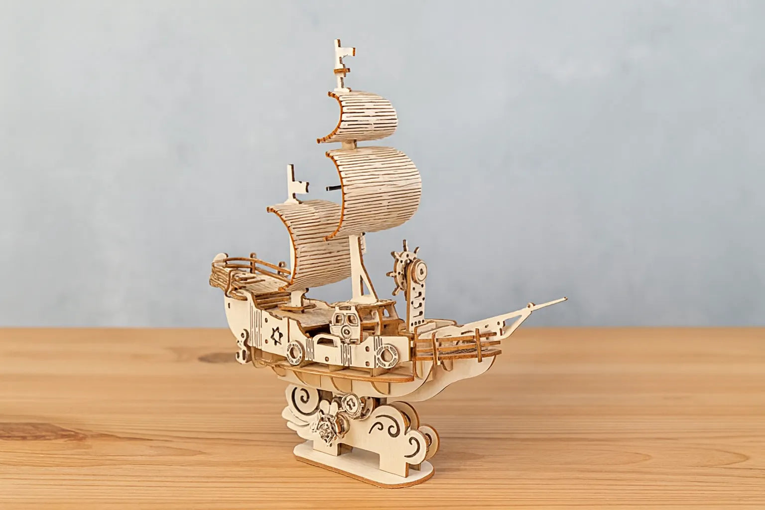Maquette en bois d’un navire des vents stylisé, ornée de voiles courbées et de roues décoratives, posée sur une table en bois devant un mur gris clair.