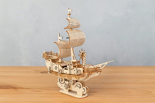 Maquette en bois d’un navire des vents stylisé, ornée de voiles courbées et de roues décoratives, posée sur une table en bois devant un mur gris clair.