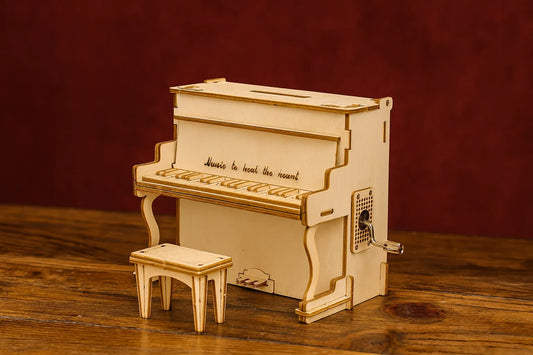 Maquette en bois d’un piano droit avec boîte à musique et tabouret, posée sur une table en bois devant un fond rouge foncé.