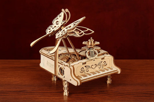 Maquette en bois d’un piano à queue miniature décoré de papillons finement découpés, présentée sur une table en bois devant un rideau rouge profond.