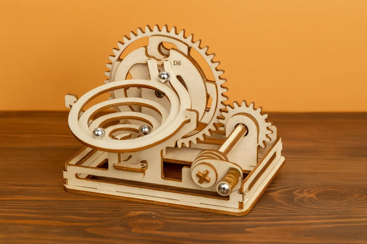 Maquette en bois d’une spirale de billes, avec engrenages circulaires et billes métalliques argentées, présentée sur une table en bois devant un mur orangé.