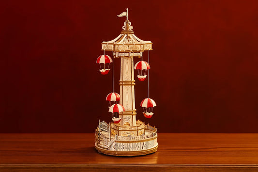 Maquette en bois d’une tour de parachutes avec nacelles rouges posée sur une table en bois devant un mur rouge.