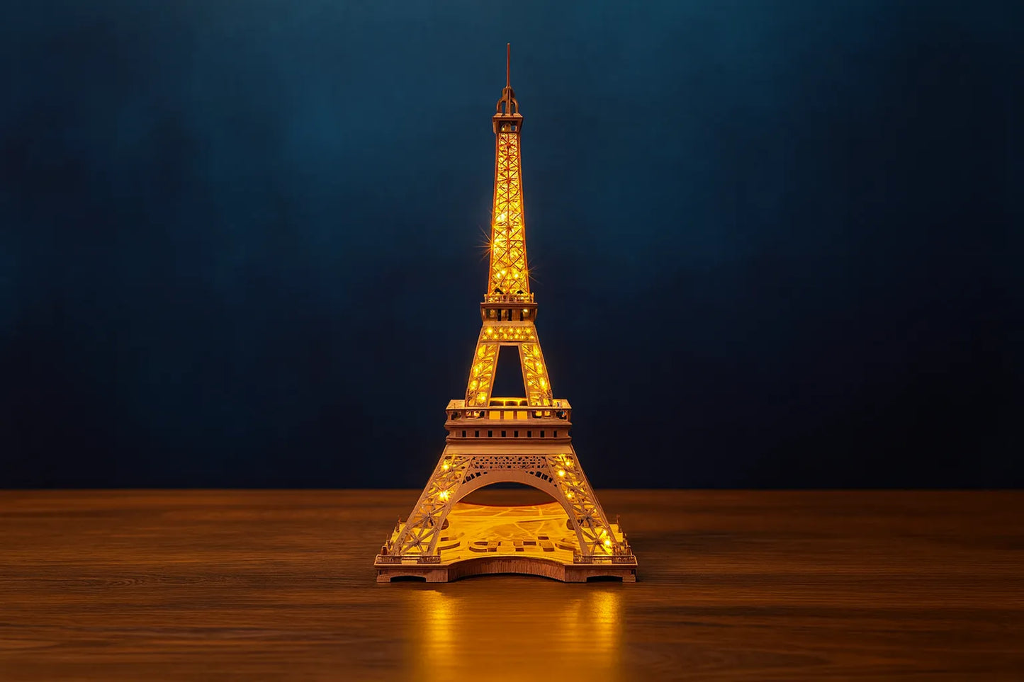 Maquette en bois illuminée de la tour Eiffel, posée sur une table en bois devant un fond bleu foncé.