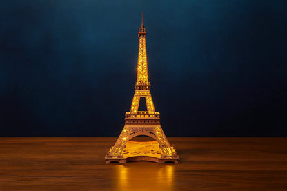 Maquette en bois illuminée de la tour Eiffel, posée sur une table en bois devant un fond bleu foncé.