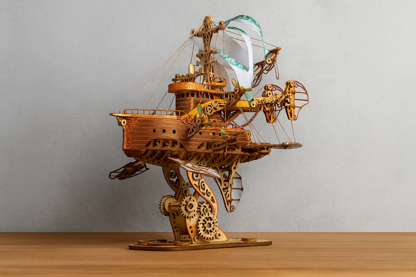 Maquette en bois d’un vaisseau steampunk détaillé, doté de voiles blanches et de structures mécaniques, posée sur une table en bois devant un mur gris clair.
