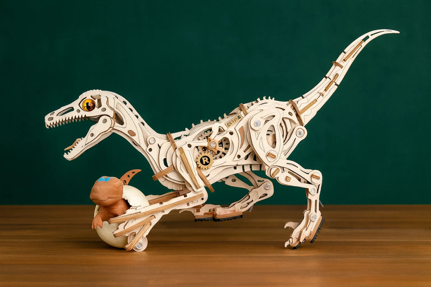 Maquette en bois articulée d’un vélociraptor avec engrenages visibles et un bébé dinosaure dans un œuf, posée sur une table en bois devant un fond vert foncé.
