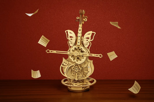 Maquette en bois d’un violoncelle décoré d’ailes de papillons et de motifs féeriques, entourée de partitions flottantes sur une table en bois avec un fond rouge artistique.
