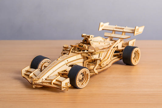Maquette en bois d’une voiture de Formule 1 avec pneus noirs, posée sur une table en bois devant un mur gris claire.