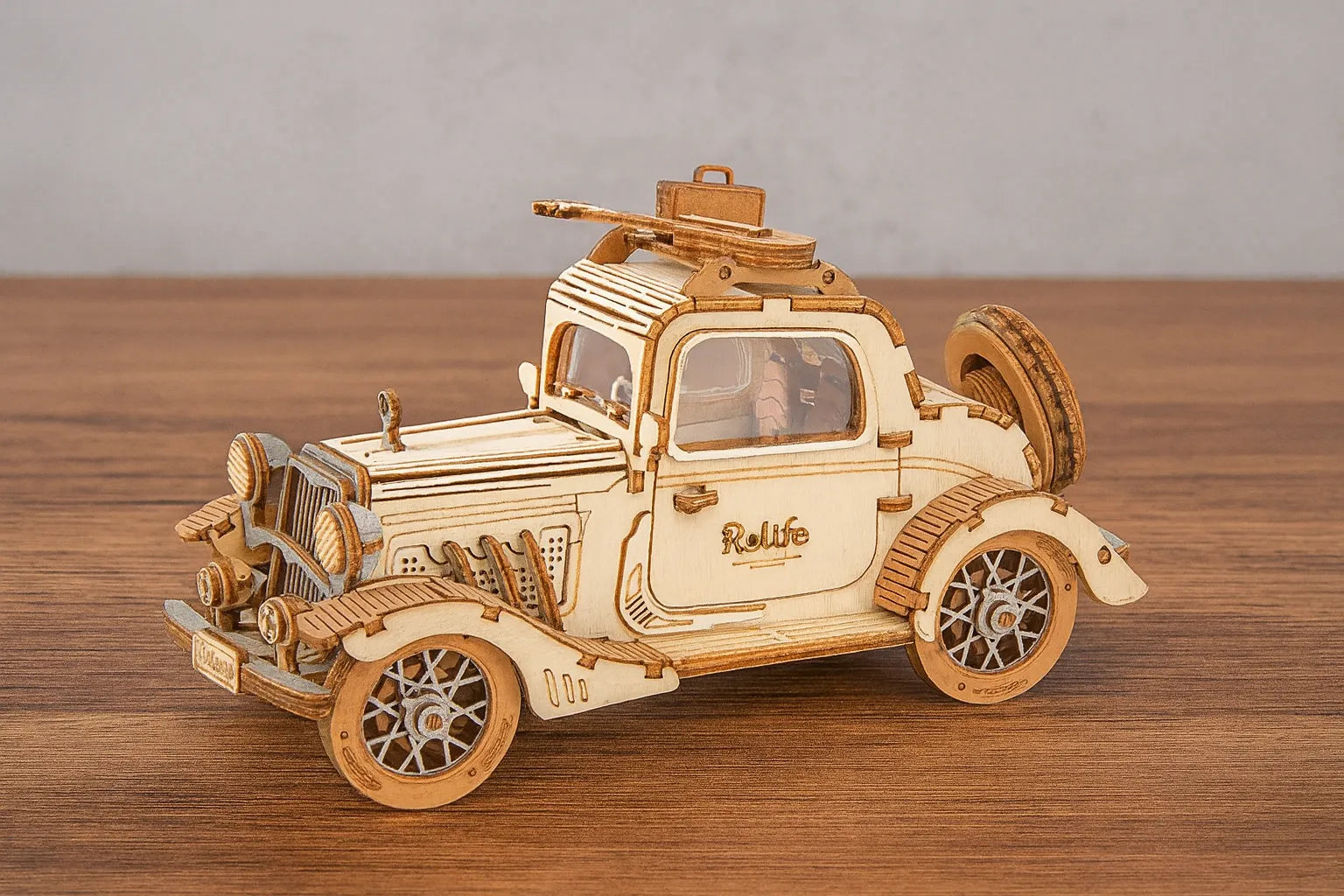 Maquette en bois d’une voiture vintage avec le logo “Rolife” sur la portière et une valise sur le toit, placée sur une table en bois devant un mur gris clair.