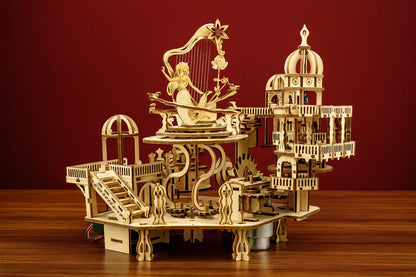 Modèle mécanique en bois de « Le jardin magique », mettant en scène une sirène harpiste entourée de structures miniatures et d’escaliers détaillés, posé sur une table devant un fond rouge.